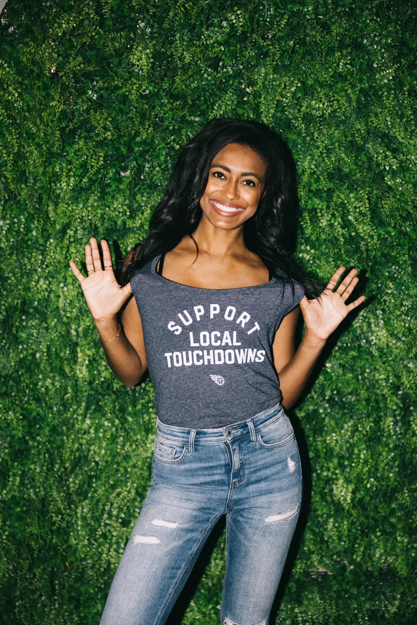 A woman in a Support Local Touchdowns tee smiling against a green hedge wall